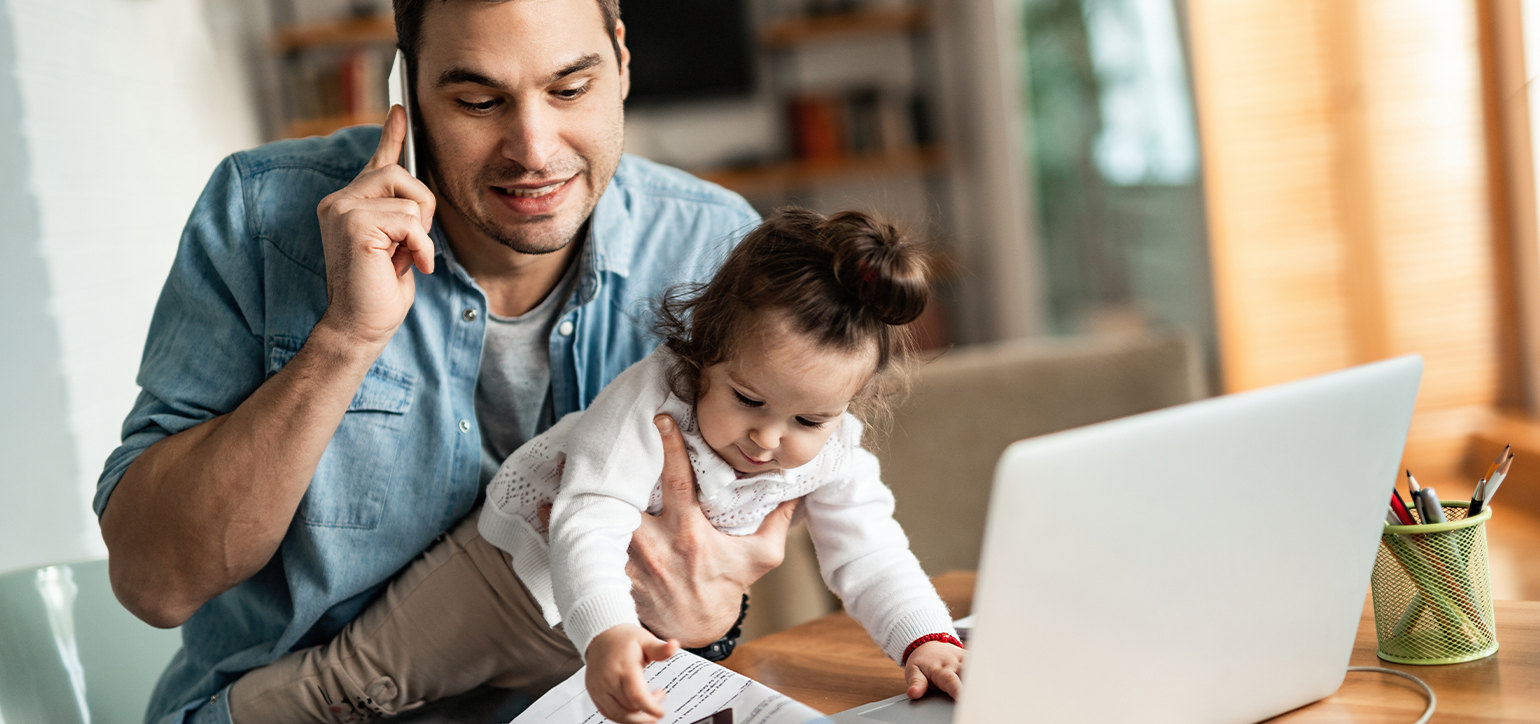 Man holding a child whilst speaking on a telephone and sitting in front of a laptop. 