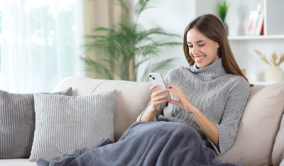 Happy woman using phone covered by blanket sitting on a couch at home